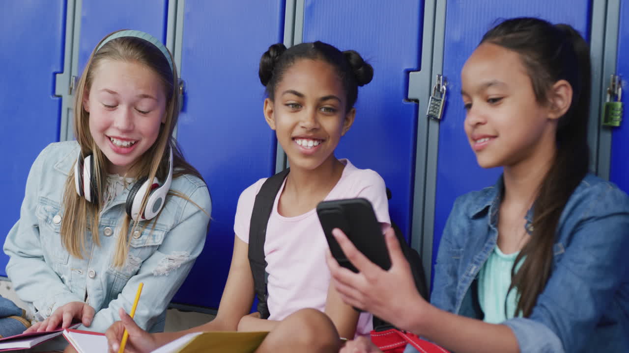 Video of three happy, diverse schoolgirls looking at smartphone, sitting by lockers, copy space