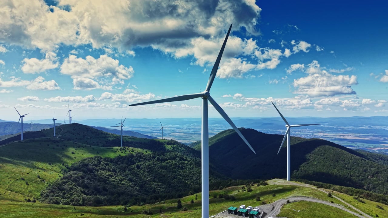 Aerogeneradores se alzan sobre colinas verdes bajo un cielo azul despejado con nubes al fondo en un sitio de energía renovable en un día soleado.