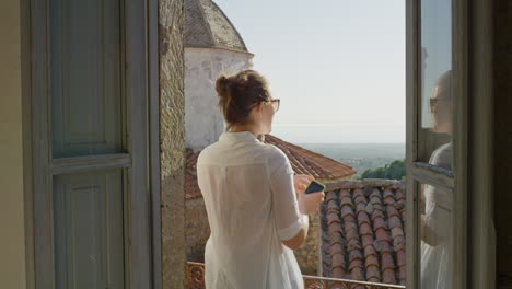 mujer hermosa usando un teléfono inteligente navegando en línea enviando mensajes de texto disfrutando de compartir la experiencia de las vacaciones de verano relajándose en el balcón de una villa rústica usando gafas de sol