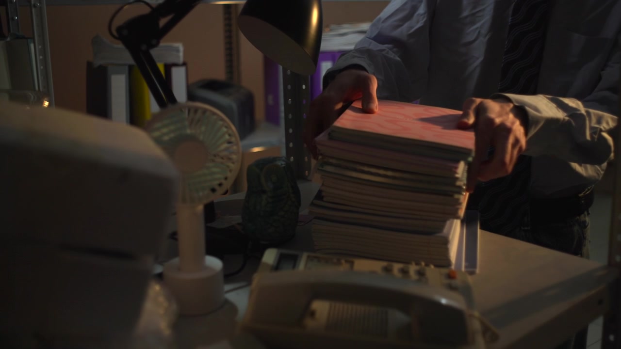 Un ambiente di lavoro d'ufficio disordinato con cartelle impilate, carte sparse e un piccolo ventilatore sulla scrivania, con raccoglitori e scaffalature visibili sullo sfondo sotto una luce.