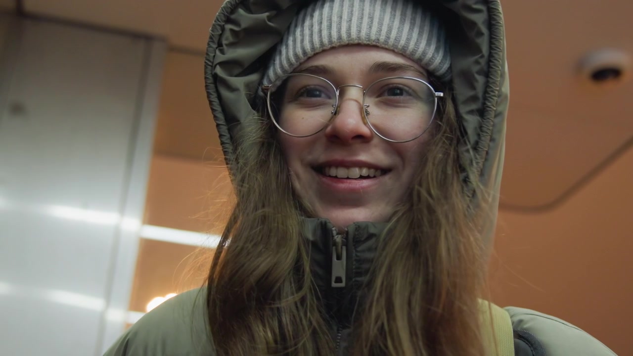 Young white woman in puffer jacket beanie, candid closeup in metro station with glasses and soft warm lighting, visible breath and grainy cinematic texture, subtle smile and thoughtful gaze, winter