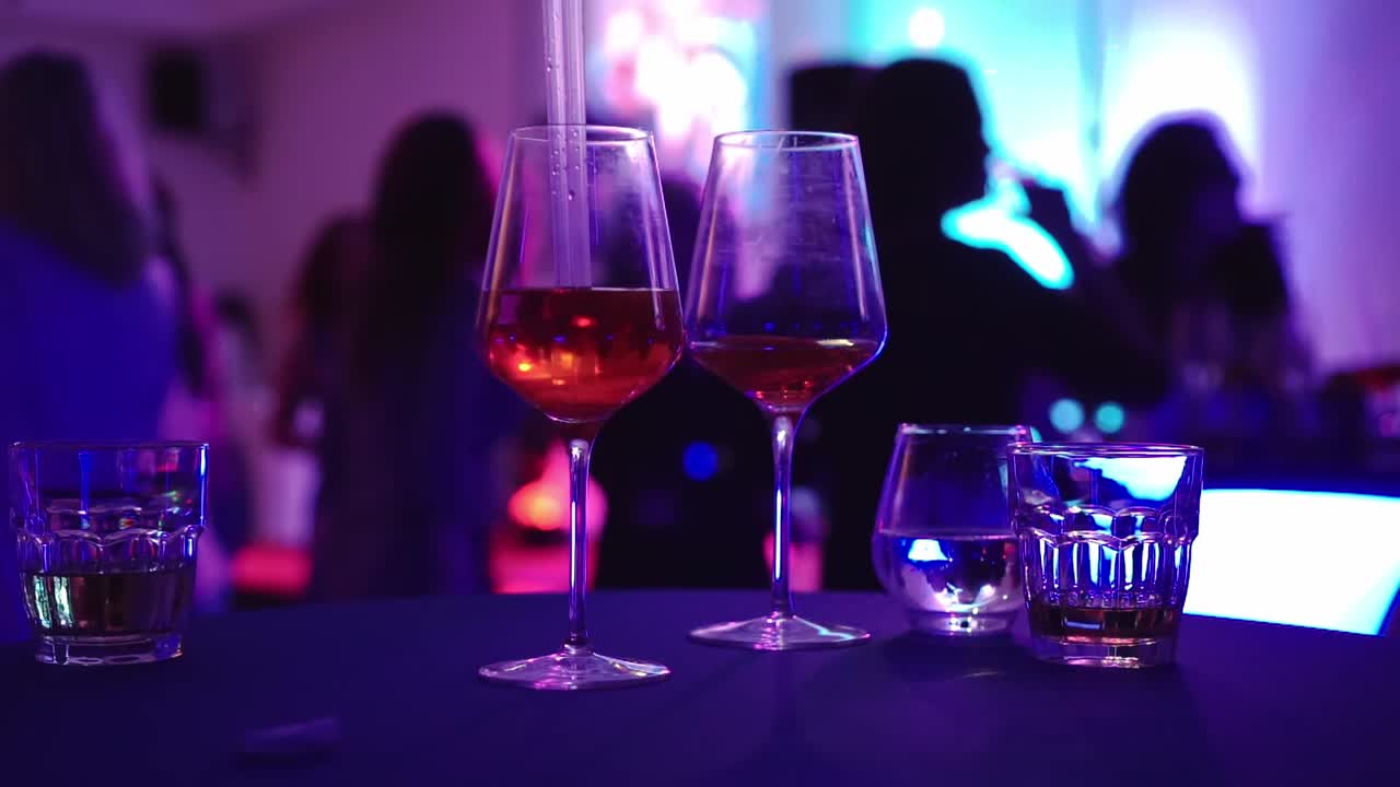 Copos com álcool na mesa e festa no plano de fundo, close em câmera lenta