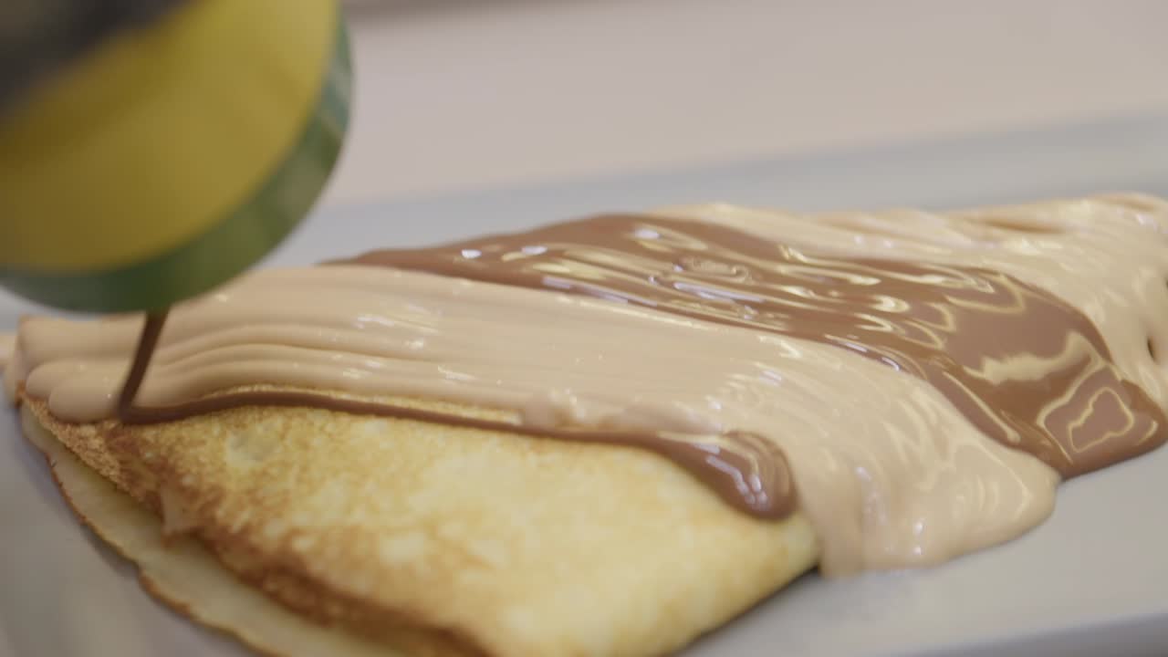 A close-up shot of rich, smooth chocolate and white cream being poured from a squeeze bottle over a freshly made crepe or pastry