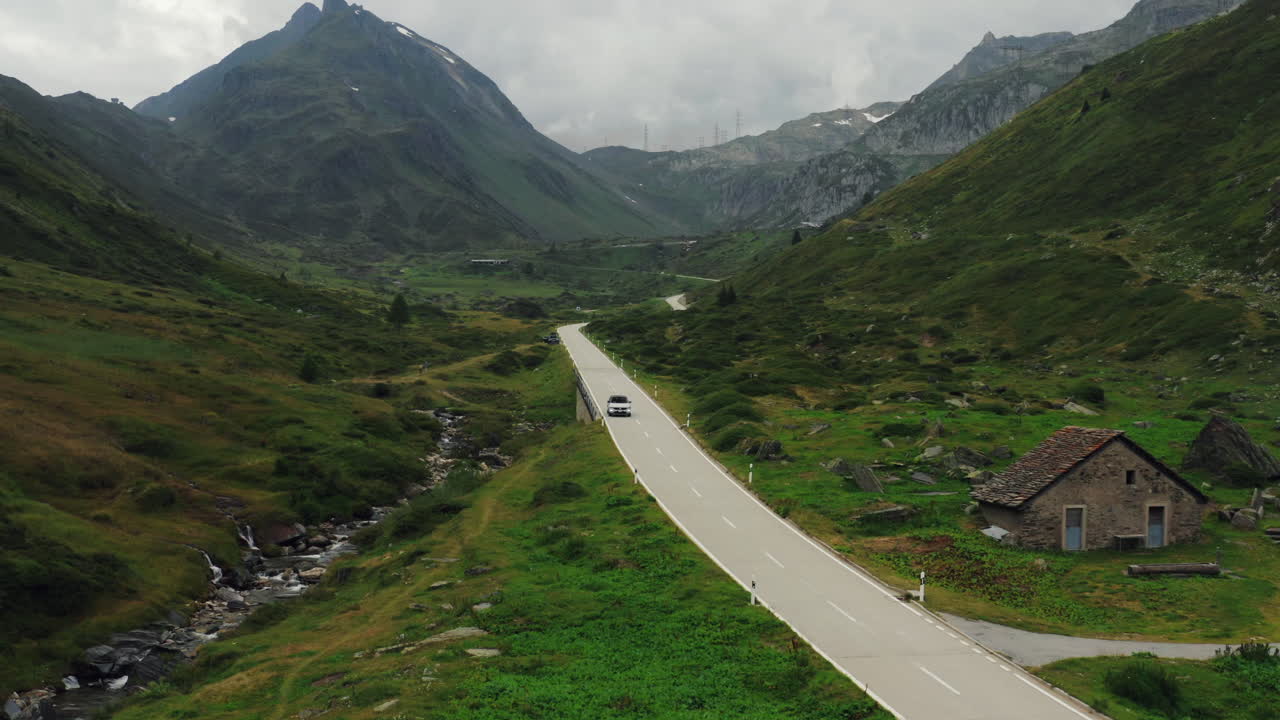 Aerial wide shot - white car passing a stone cottage on narrow mountain road in alpine valley under a cloudy sky. Switzerland.