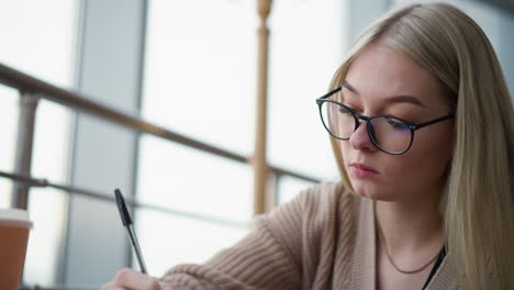 close-up of young lady with glasses focusing intently while writing with a pen on paper, demonstrating concentration in a quiet workspace or study session, with blurred background