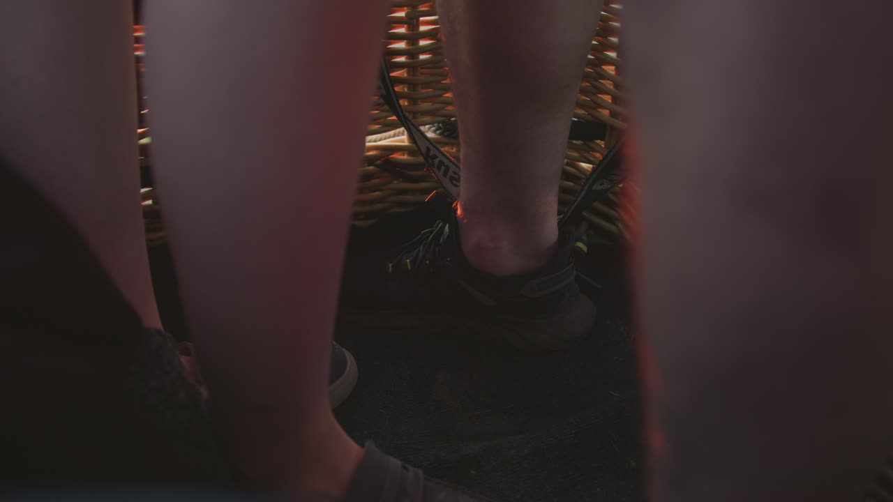 close up legs of couple standing in wicker balloon basket illuminated by warm sunset light with safety straps visible as they float above sprawling fields at golden dusk