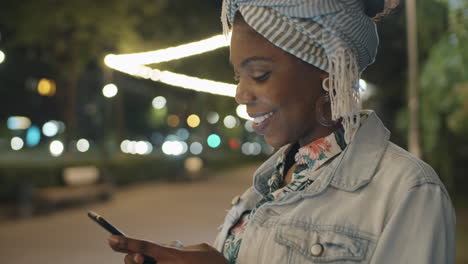 Mujer-Afroamericana-Sonriendo-Y-Enviando-Mensajes-De-Texto-Por-Teléfono-En-El-Parque-Por-La-Noche