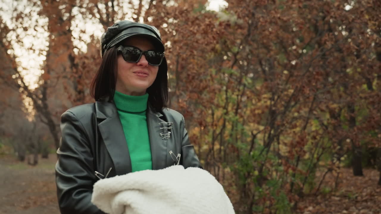 Donna bianca che sorride all'aperto tenendo una coperta in un parco panoramico, indossa una giacca di pelle, un dolcevita verde, un berretto borchiato e occhiali da sole oversize, gesti giocosi, movimento arioso, toni caldi cinematografici