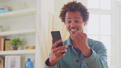 Celebrando-Al-Hombre-En-Casa-Usando-Un-Teléfono-Móvil-Recibiendo-Buenas-Noticias-Sobre-Una-Nueva-Empresa-Comercial