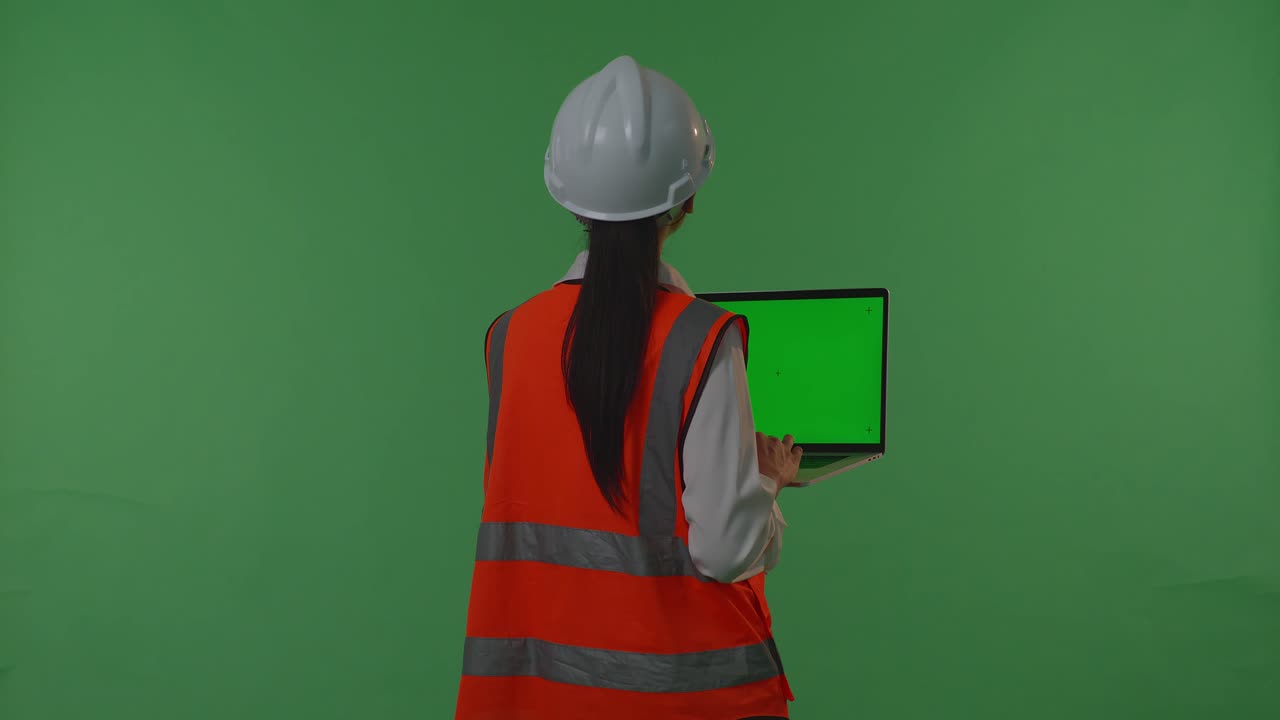 안전 헬을 입은 아시아 여성 엔지니어가 녹색 스크린 노트북에서 작업하고, 해가 지거나 해가 뜨는 시간 동안 녹색 화면 배경 스튜디오에 서있는 동안 주변을 둘러보고 있습니다.