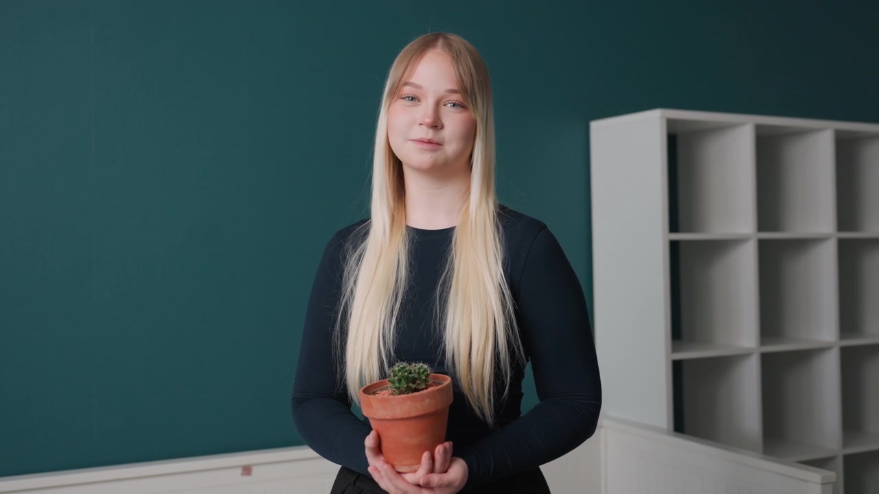 Bílá žena drží rostlinu v bytě, nadějná nově příchozí aranžuje terakotový květináč u prázdných polic, tyrkysová stěna v pozadí, dlouhé blond vlasy, ležérní černý svetr, jemné denní světlo, minimalistický interiér.
