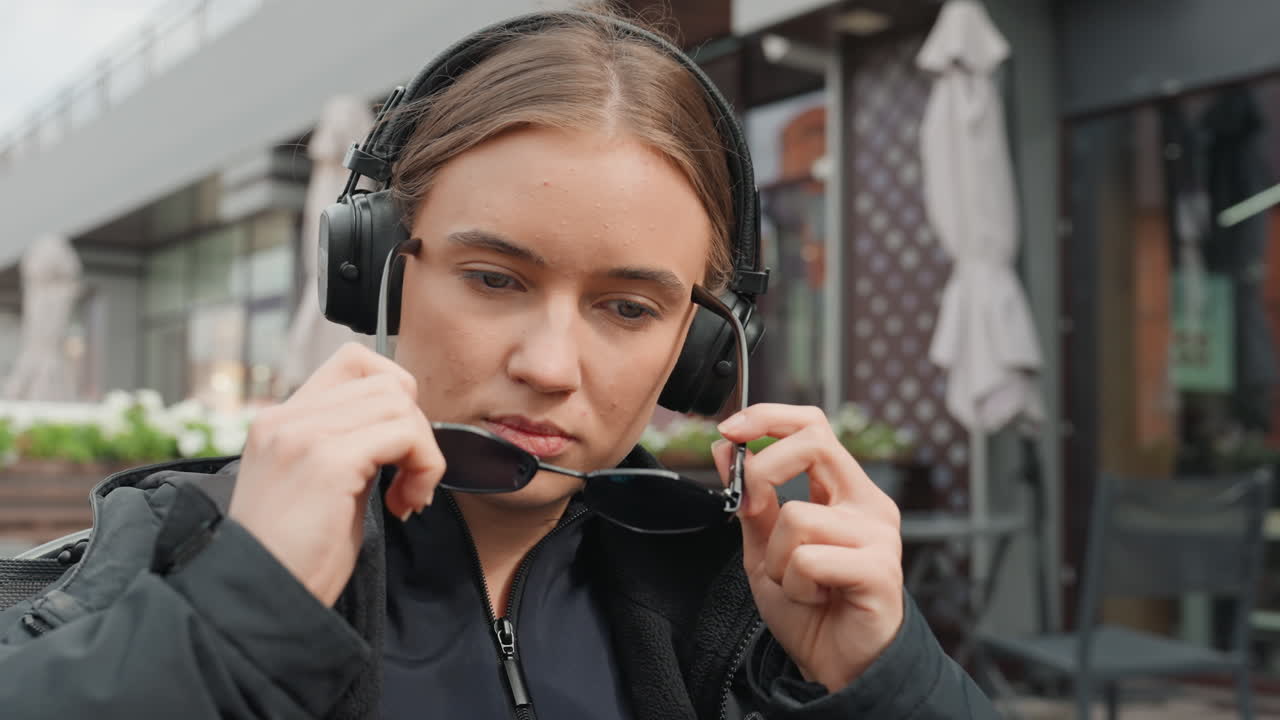Kaukasisk kvinne på kafé med hodetelefoner, som roter i ryggsekken, justerer solbriller, nipper til kaffe fra take away-kopp, flettet hår synlig, fokusert uttrykk, urbant terrassemiljø
