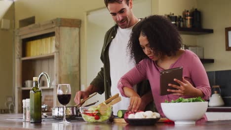 happy diverse couple preparing a meal together in kitchen, using recipe on tablet