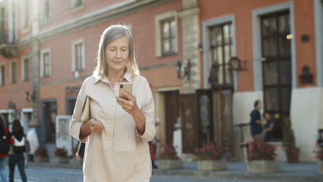 Senior-Gut-Aussehende-Dame,-Die-In-Der-Altstadt-Auf-Der-Straße-Spazieren-Geht-Und-Auf-Das-Smartphone-Tippt