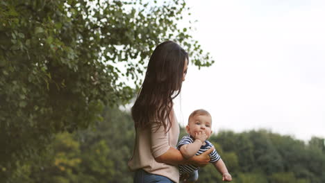 Feliz-Joven-Madre-Sosteniendo-A-Su-Lindo-Bebé-En-El-Brazo-Balanceándose-Como-Un-Avión-Al-Aire-Libre