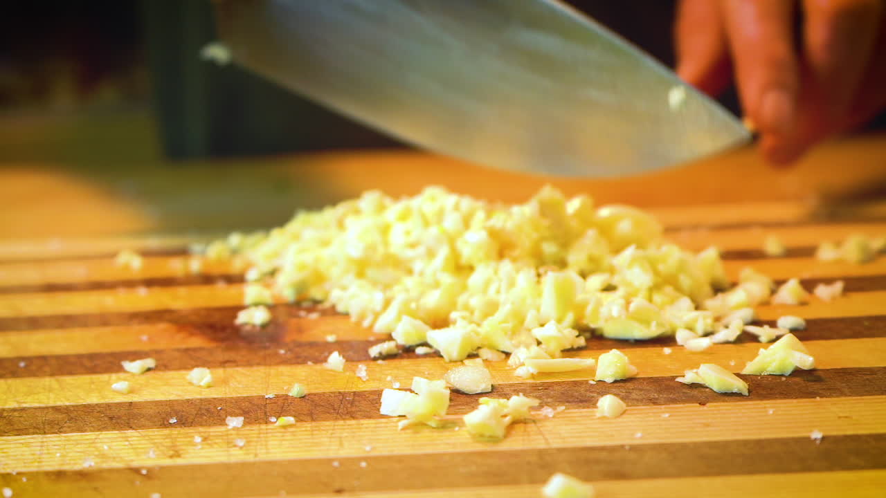 Garlic is being minced with a chef's knife on a striped wooden cutting board