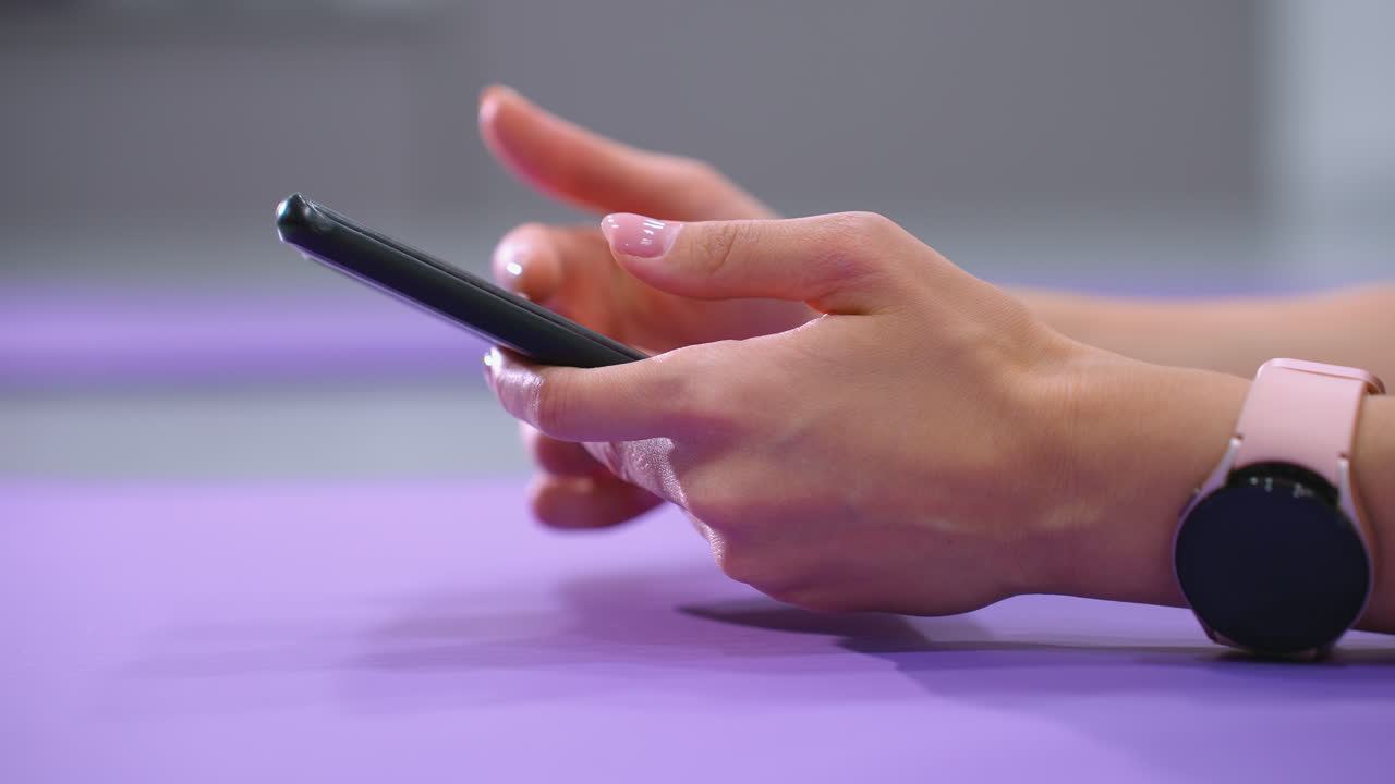 nahaufnahme von händen, die durch das smartphone gleiten, während sie auf einer lila yoga-matte liegen. die person trägt eine smartwatch und hat gut gepflegte nägel. weiches beleuchtung verbessert die verwendung moderner technologie.