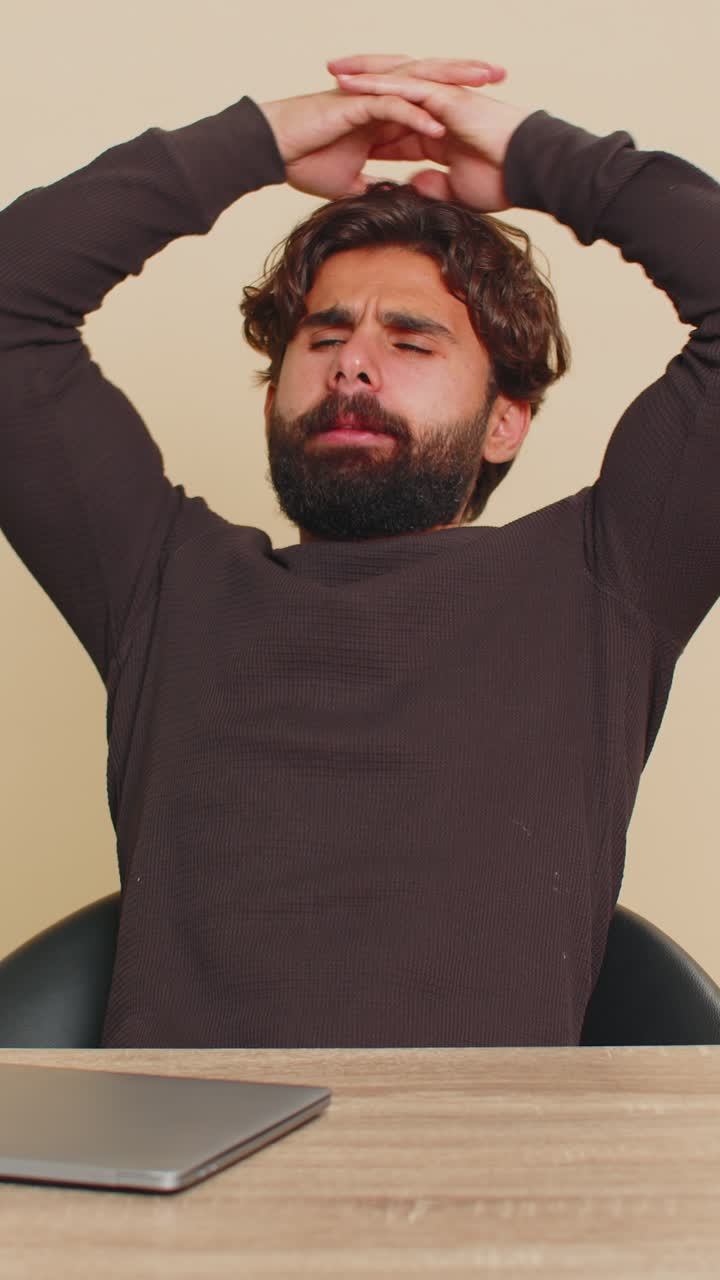 Bored indian young man freelancer closing laptop and sitting at table against beige background