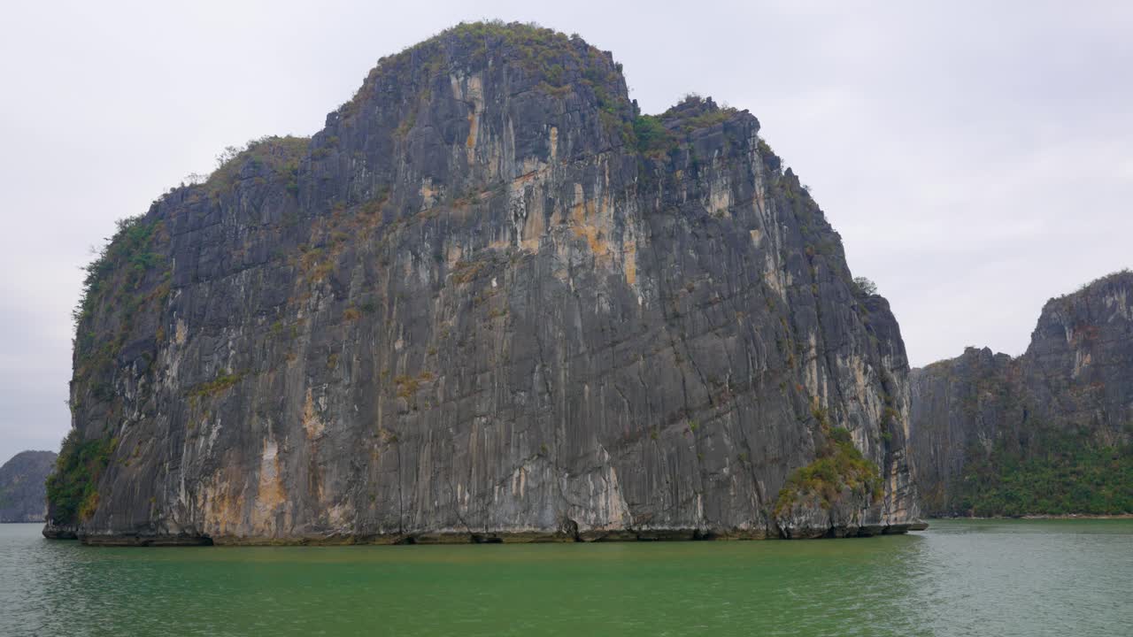 Towering limestone karst formation rises dramatically from the emerald-green waters of Ha Long Bay, its weathered gray cliffs stretching skyward while scattered vegetation clings to its rocky facade