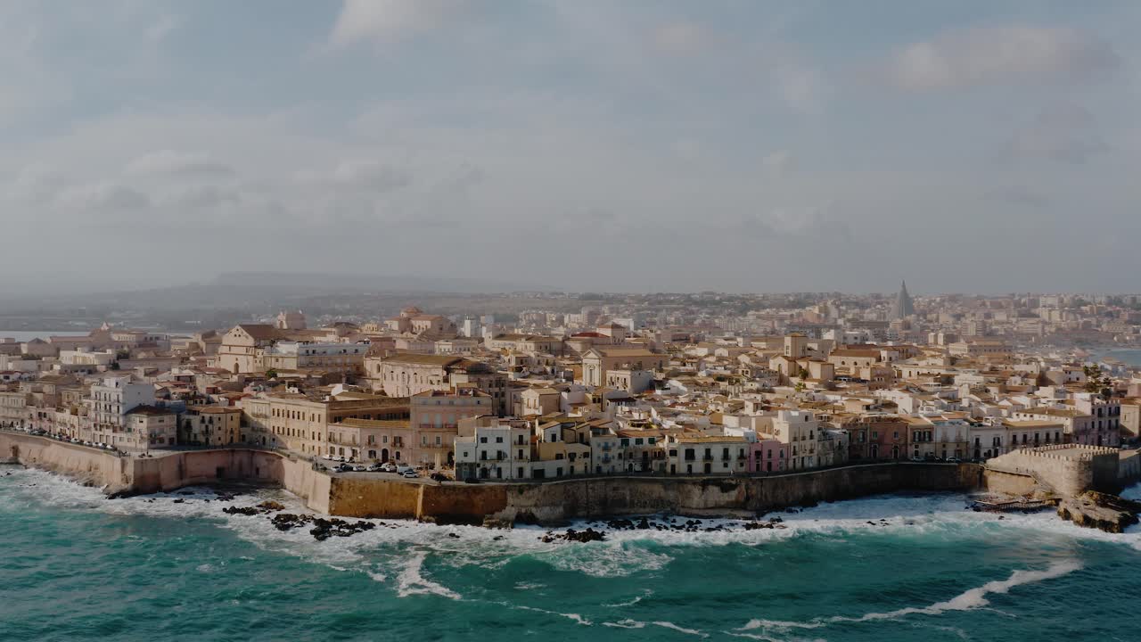 Drone flying towards the island of Ortigia in Sicily. Historic old town of Syracuse by the Mediterranean sea with large waves crashing onto the shore. UNESCO world heritage.Popular travel destination.