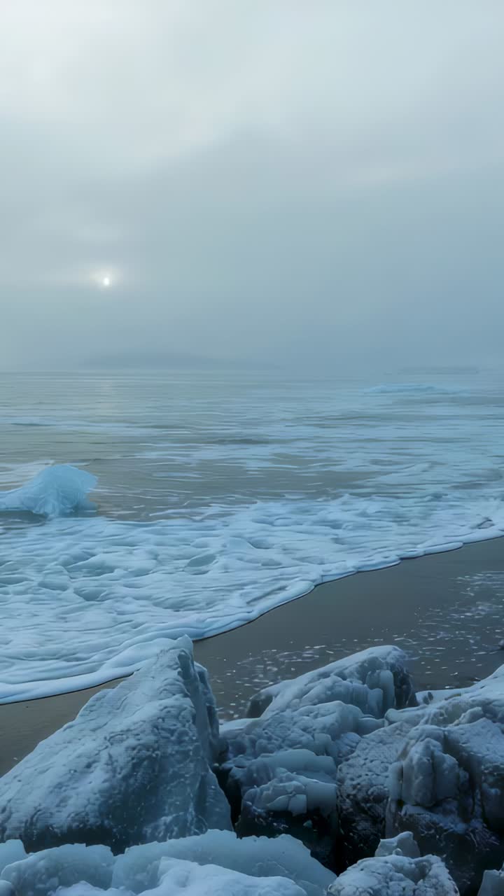 Vertical video: Breaking wave approaching, pushing foamy surf across sand and rocks under pale sun