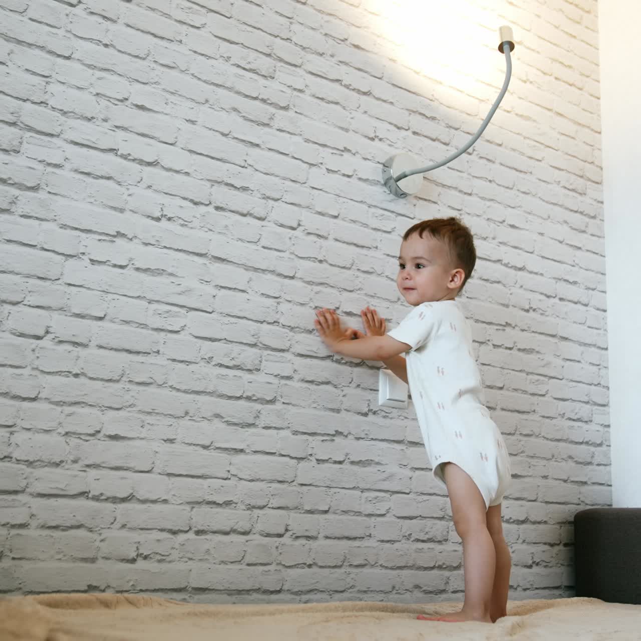 Lovely Caucasian toddler boy in white bodysuit stands on the bed. Little kid plays happily with lamp. Low angle view