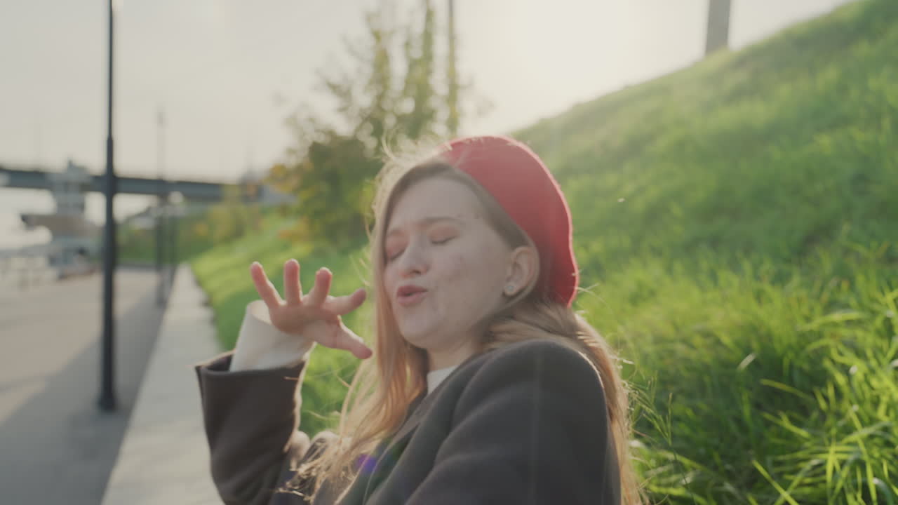 Kvinne som feirer ved en vannkantsti, danser, vinker og ler under varmt sollys. Leken håndbevegelse, grønn skråning og havnebakgrunn, stilig kåpe og beret, et bekymringsløst livsstilsøyeblikk.