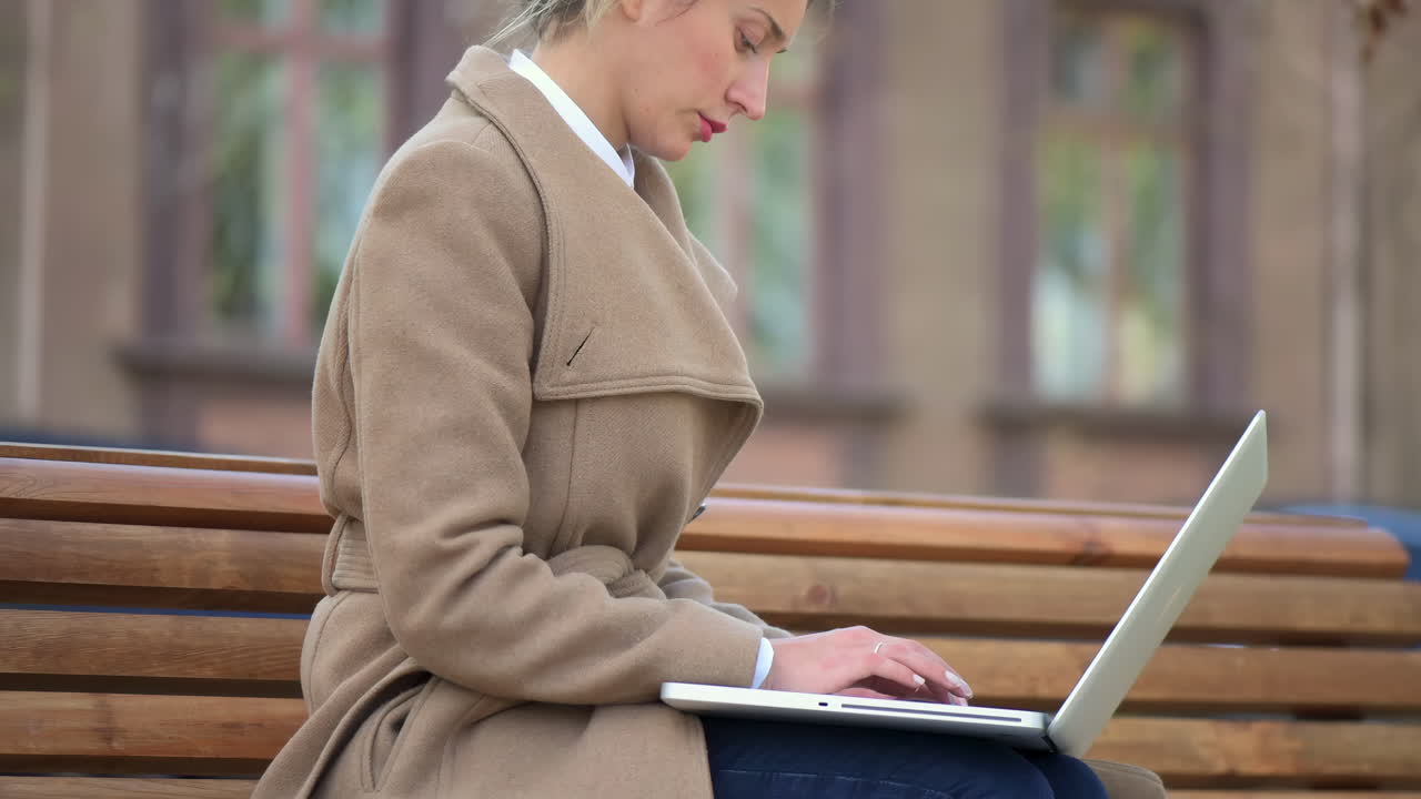 Woman in a brown coat working on her laptop on a bench in the park