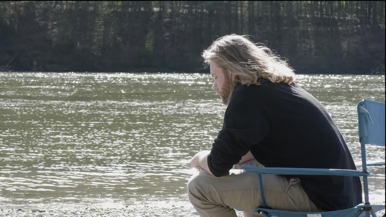 En ung man med långt blont hår sitter tankfullt i sin campingstol.