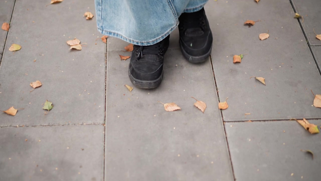 Close up of person feet in black shoes and light blue jeans walking on gray tiled pavement with scattered autumn leaves. focus remains consistently low angle capturing movement from foot