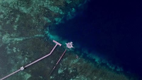 Barco-Jukung-En-Las-Islas-Raja-Ampat-Indonesia-Estacionado-En-Un-Muelle-Mientras-Otro-Barco-Más-Pequeño-Navega,-Vista-Aérea-Superior-Bajando-El-Tiro