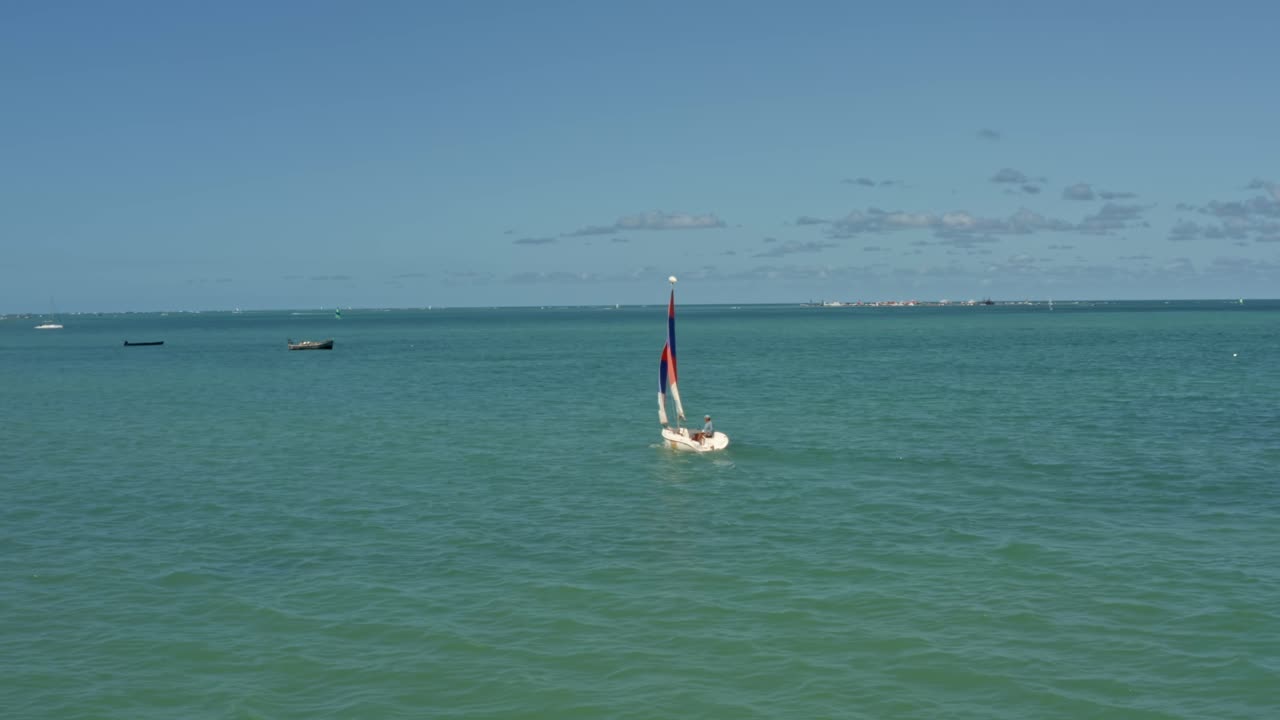 toma aérea giratoria siguiendo un colorido barco de vela cerca de la playa tropical de bessa en la capital costera de joao pessoa, paraiba, brasil con la famosa isla de arena roja en el fondo