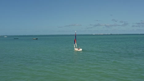 rotating aerial shot following a colorful sail boat near the tropical bessa beach in the coastal capital city of joao pessoa, paraiba, brazil with the famous red sand island in the background