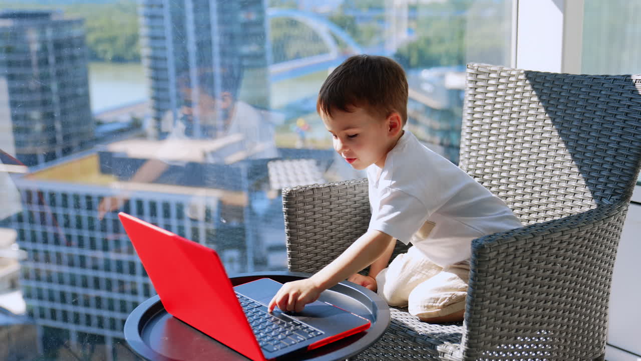 Adorable kid sitting in the chair looks focused at laptop in front of him. Smiling baby boy touches the key on the keyboard
