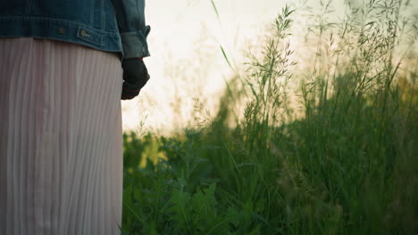 a woman in a denim jacket and pink pleated gown is walking through a tall grass field, her hand slightly brushing the plants