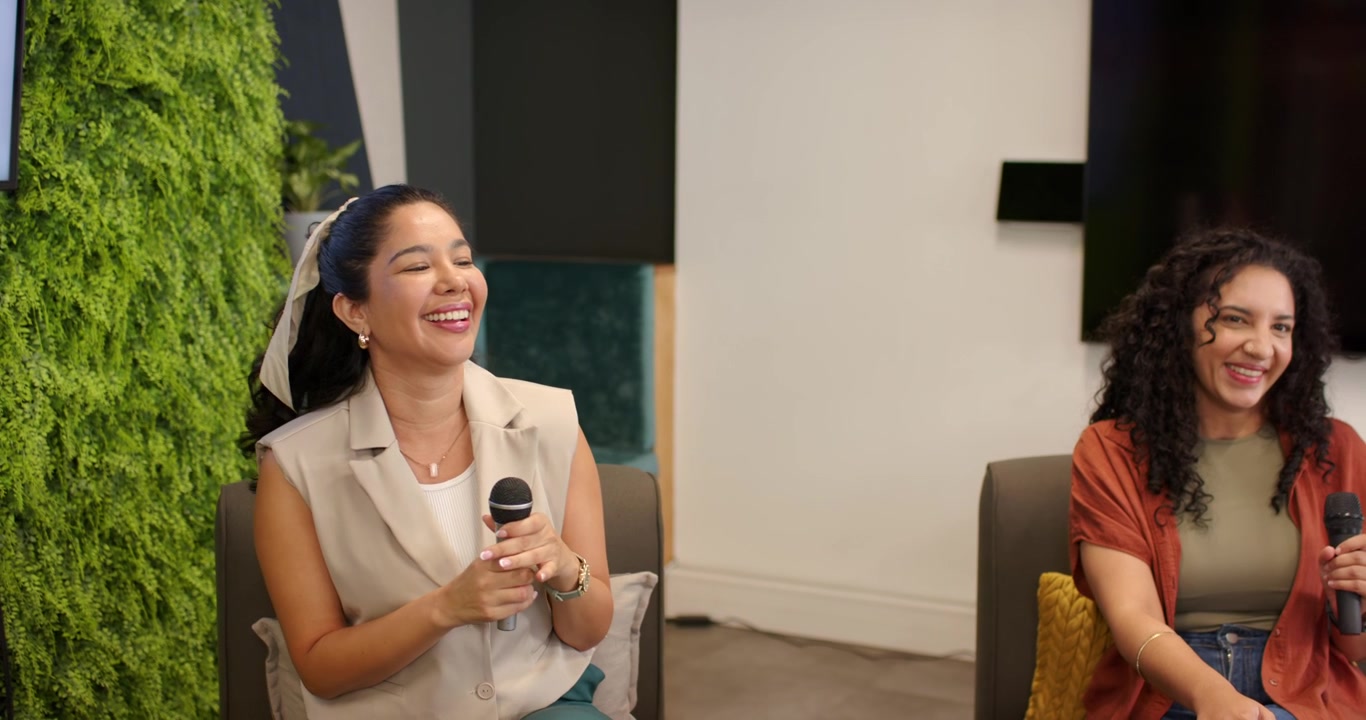 Two female speakers reacting to slide 'Let's Talk About AI', laughing with microphones in lounge