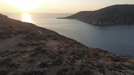 Flight-over-rocky-landscape-towards-sunset,-showing-drone-pilot-standing-on-rock,-Greece,-Kythnos-Island