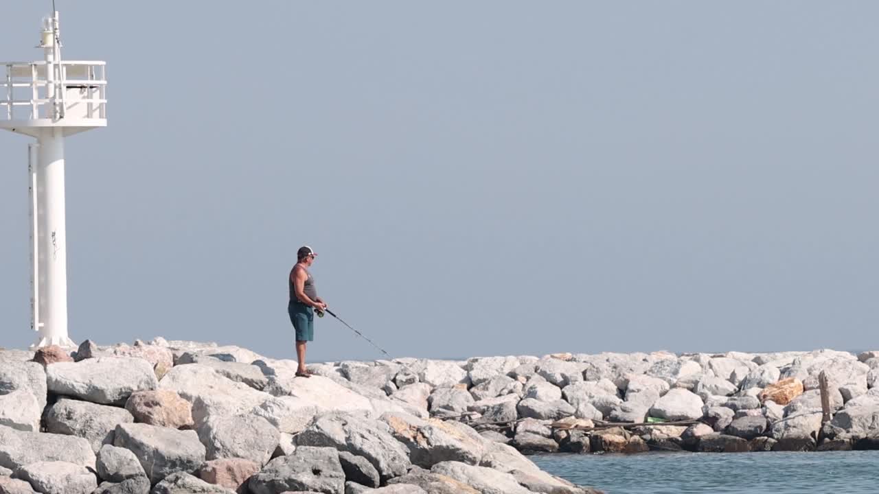 An individual fishes on a rocky breakwater beside a white tower under a clear sky.