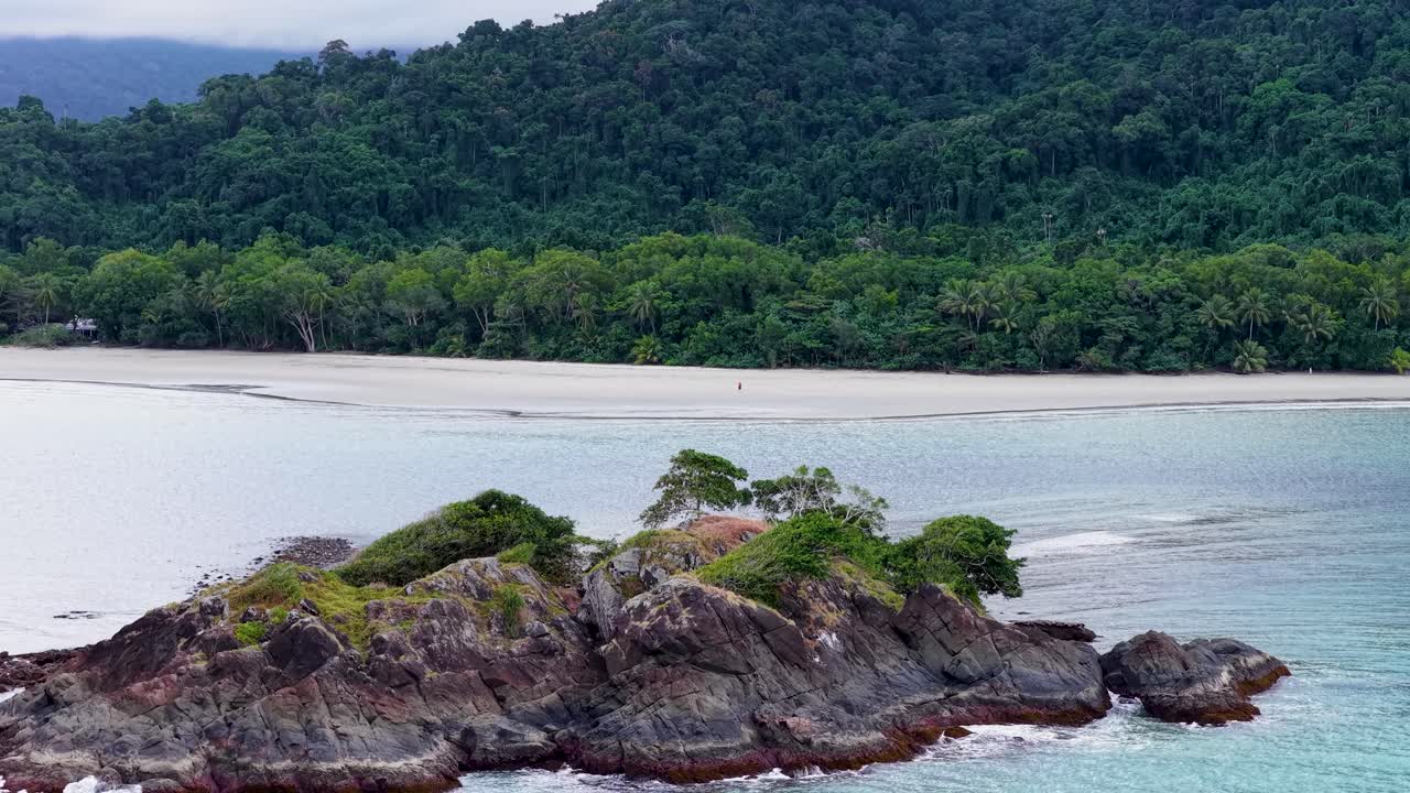 Aerial drone footage glides over a small rocky island with lush vegetation, revealing turquoise waters and dense rainforest coastline under soft, natural daylight