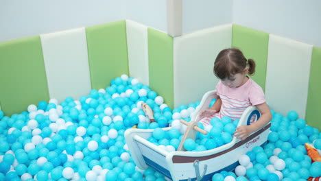 una niña pequeña juega en una piscina seca con bolas blancas y azules, sentada dentro de un bote de juguete de madera