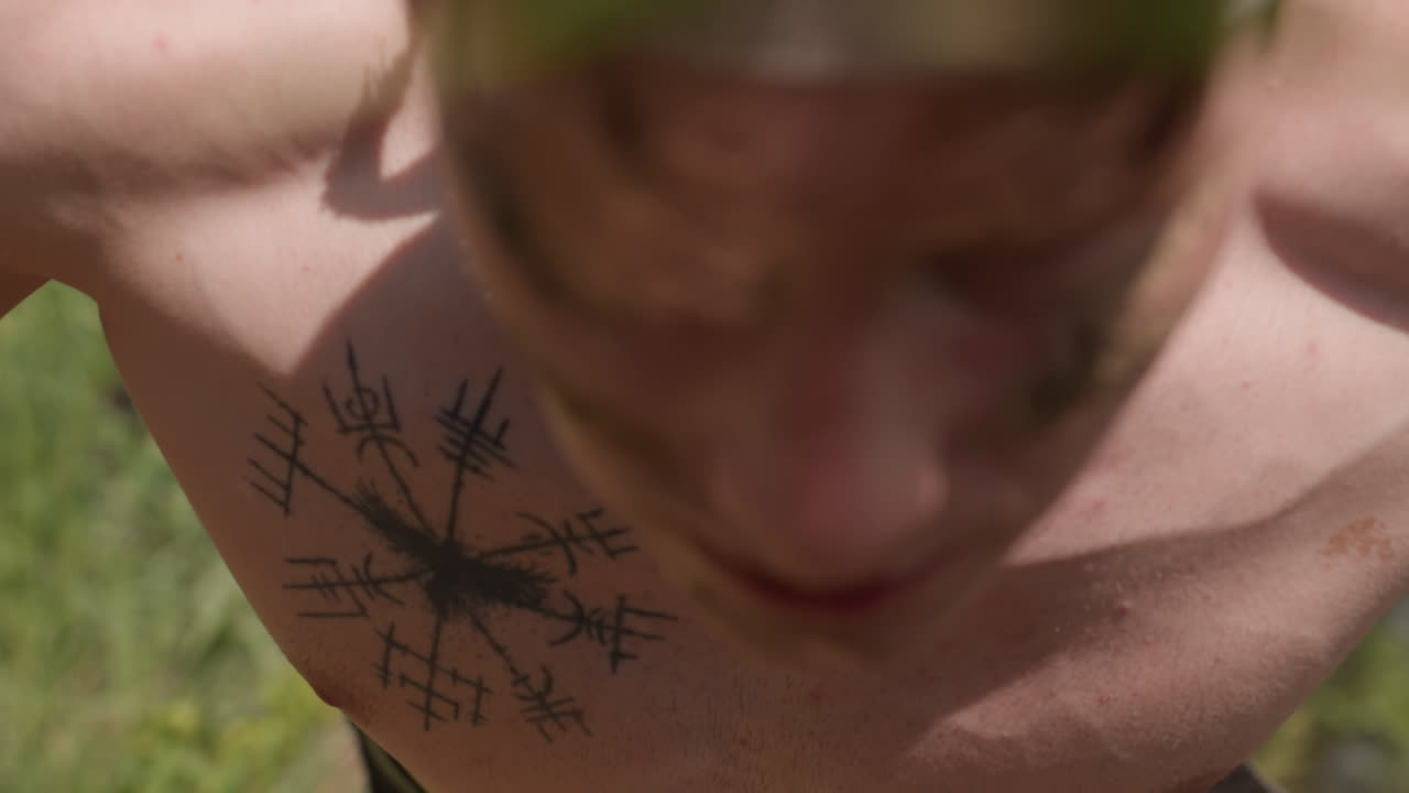 close chest view showing man eagle tattoo with arrow symbols beneath front parted hair, shirtless warrior eyeing grassy riverbank while preparing for push up exercise against lush nature backdrop