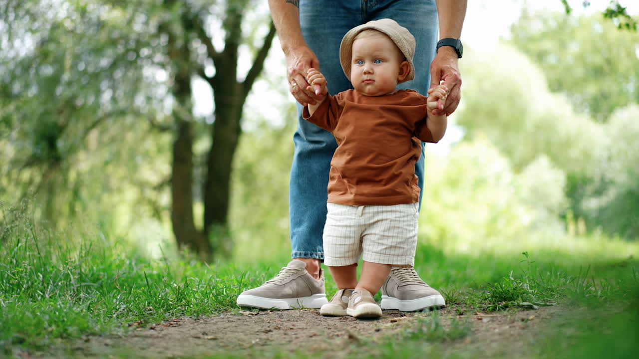Little Caucasian toddler wearing a brown t-shirt, shorts and a cap stands outdoors held by the hands. Lovely baby learning to walk supported by his father.