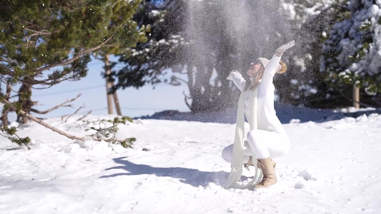Verspielte Frau mittleren Alters in warmer Kleidung wirft im Winter in einem verschneiten Wald Schnee hoch.