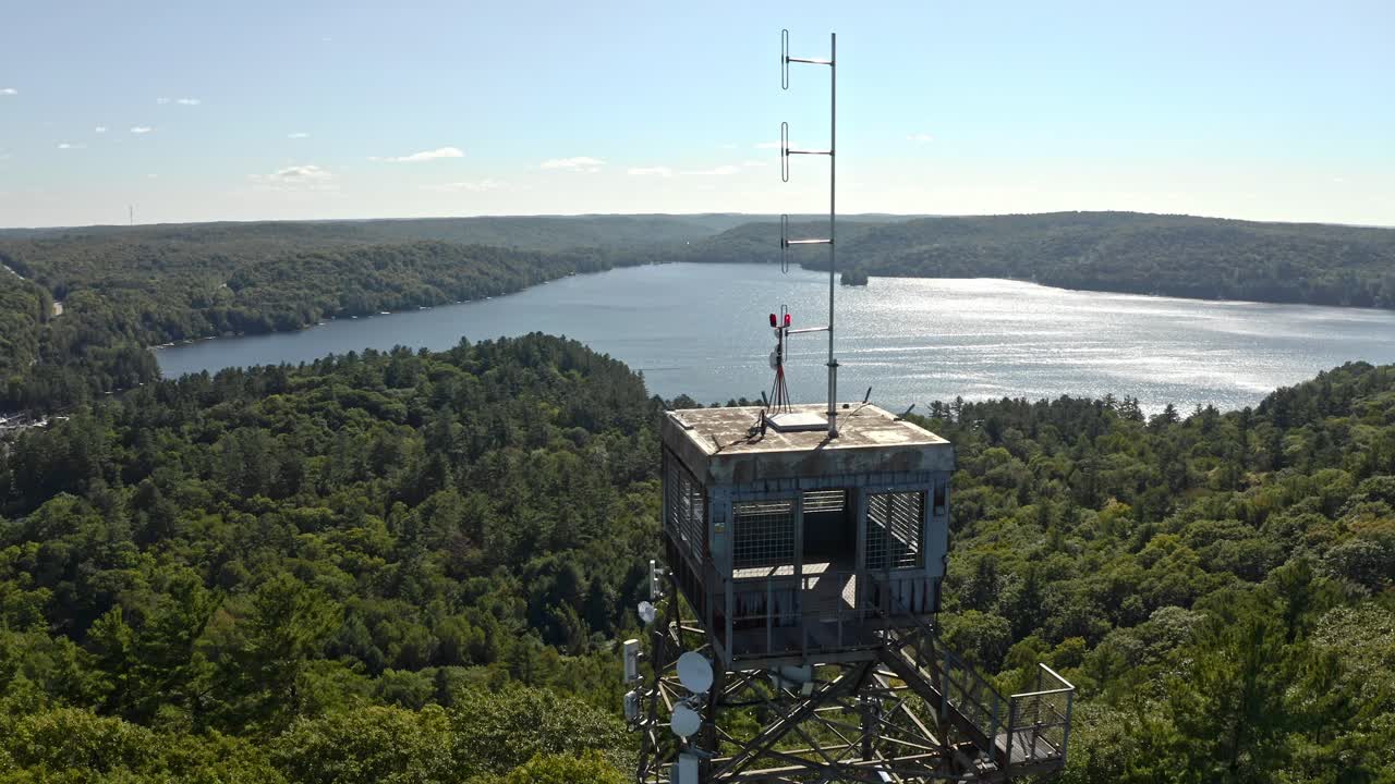 Rusty lookout tower with antenna and cell equipment on hill overlooking lake with shimmering reflections