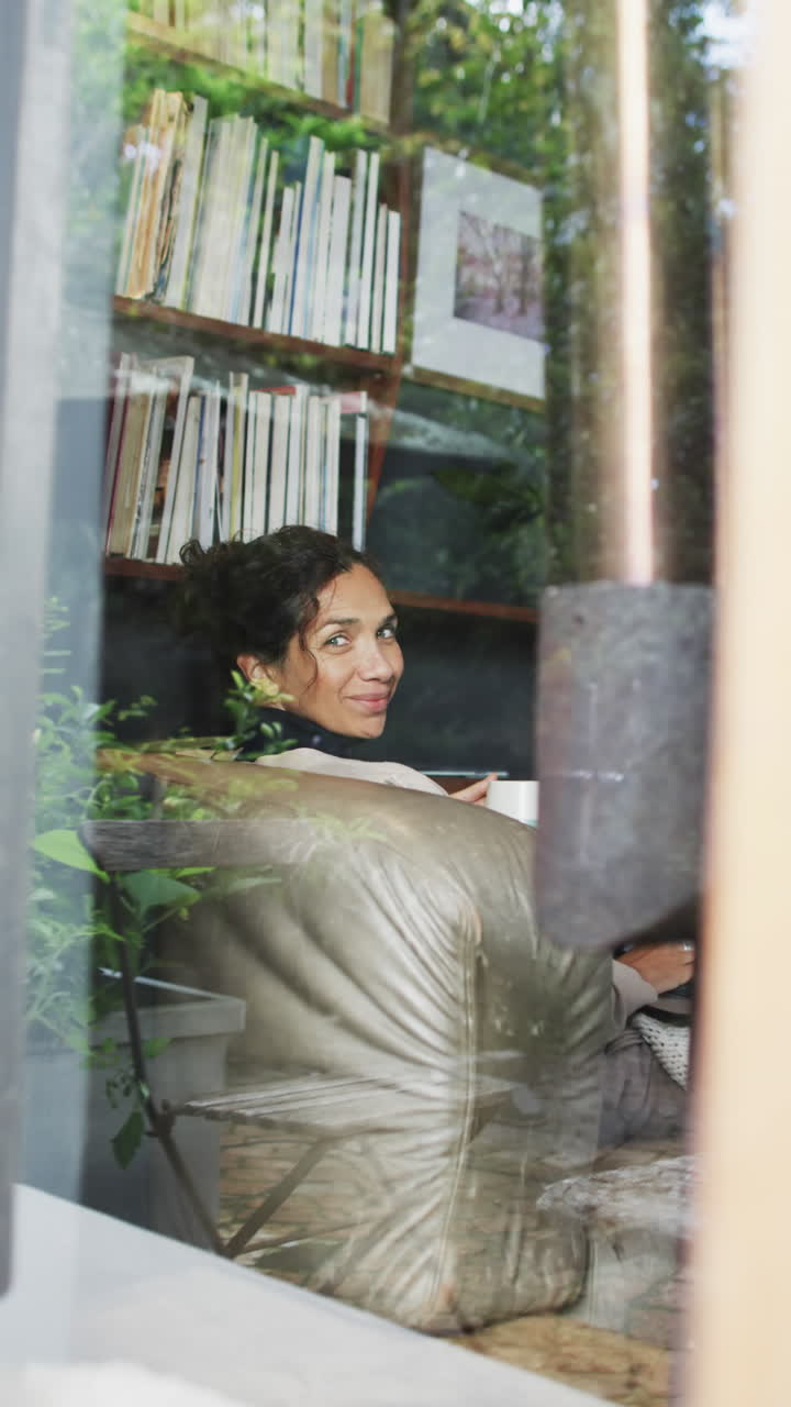 Vertical video of biracial woman lying on sofa and looking out window at home, slow motion