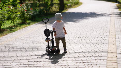 small toddler boy stands near the bicycle on the park alley. kid leaves his scooter and runs away.