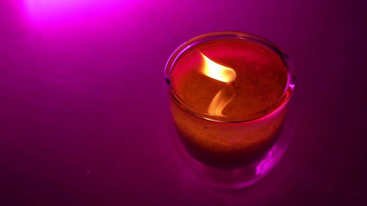 Un primo piano dall'alto di una fiamma che brucia all'interno di un portacandele di vetro trasparente. Il caldo fuoco giallo si riflette nella cera liquida, illuminato da una vivace illuminazione da studio viola su una superficie piana