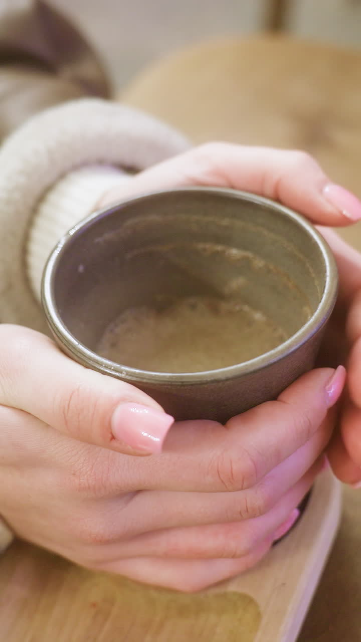 Close-up shot of woman's hand in brown shearling jacket, holding a cup of coffee, gently lifting it at cozy cafe. Warm ambiance and soft lighting create relaxing atmosphere perfect for enjoying hot drink