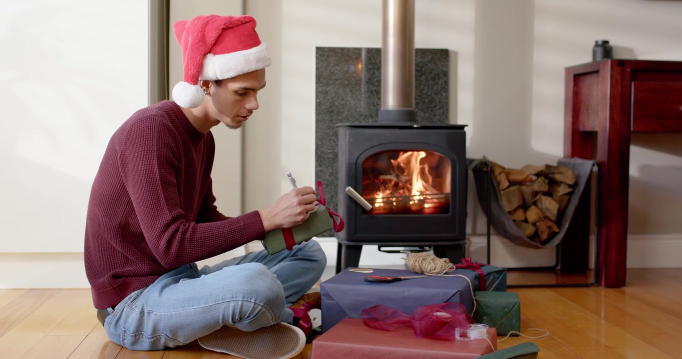 Hombre afroamericano poniéndose un gorro rojo de Papá Noel, etiquetando un regalo verde para las fiestas junto a una estufa de leña.