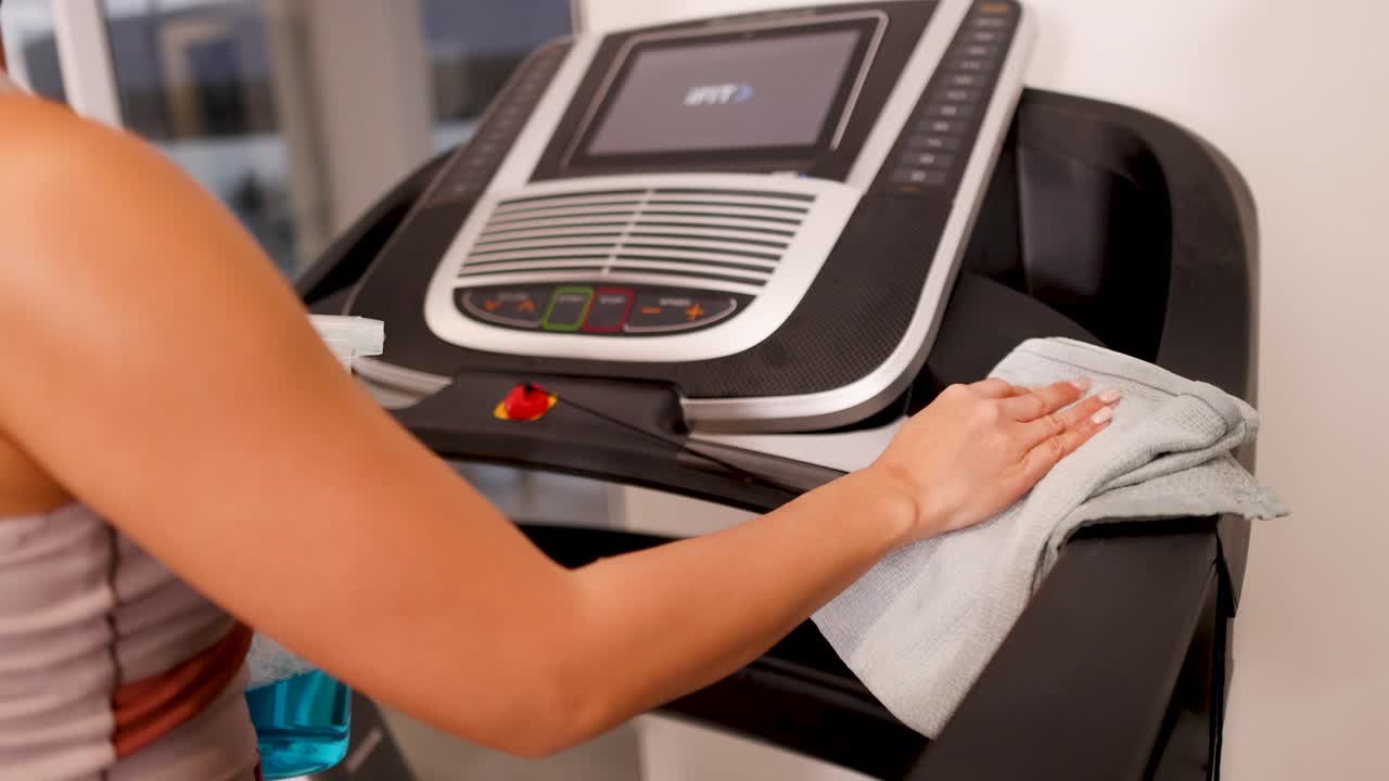 A woman cleans a treadmill with spray and cloth in a well-lit home gym, emphasizing hygiene and maintenance